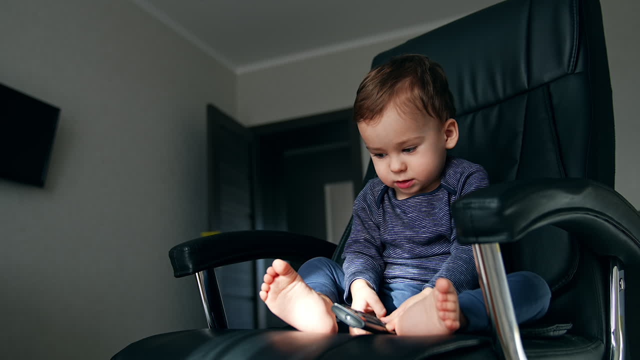 Little baby boy playing with tv remote sitting in the black office chair. Calm child with bare feet playing at home. Low angle view.