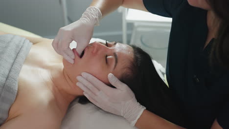 Lady lying on clinical bed opens mouth and closes eyes gently as massage therapist wearing protective gloves dips fingernail inside mouth and uses hand to massage cheeks for gentle care