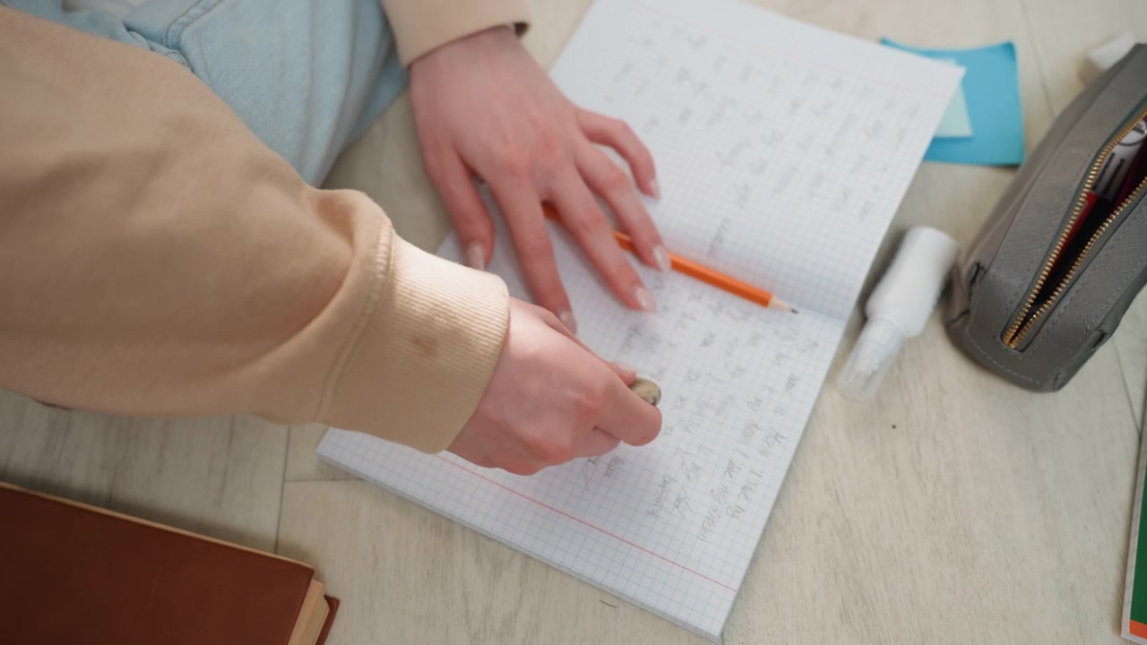 Mani che scrivono su un quaderno con la matita. Uno studente organizza appunti di studio su carta a quadretti. Accogliente scrivania di casa con braccia coperte da un maglione, astuccio con cerniera e post-it nelle vicinanze. La luce solare naturale illumina la scrittura.