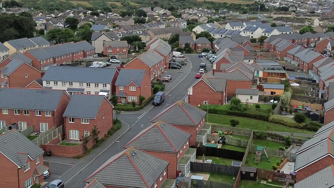 Still aerial shot pans right to left over contemporary Welsh town rooftops, with uniform pattern two-story houses. Filmed under muted afternoon skies. Ideal for urban or location content