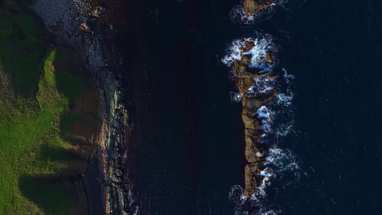 Waves gently lap against rocky shore as sunlight sparkles on the water near a green hillside at midday