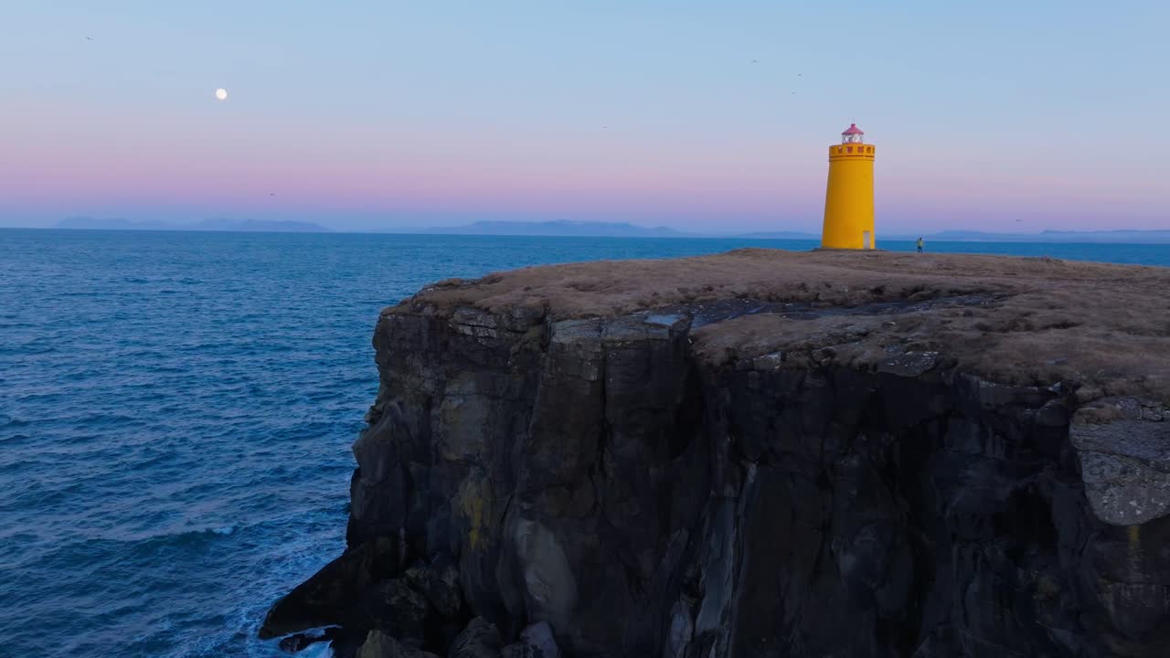 Ripresa aerea circolare di una persona solitaria che passeggia vicino a un faro su scogliere frastagliate, sfumature rosa del tramonto sullo sfondo. Ripresa aerea cinematografica