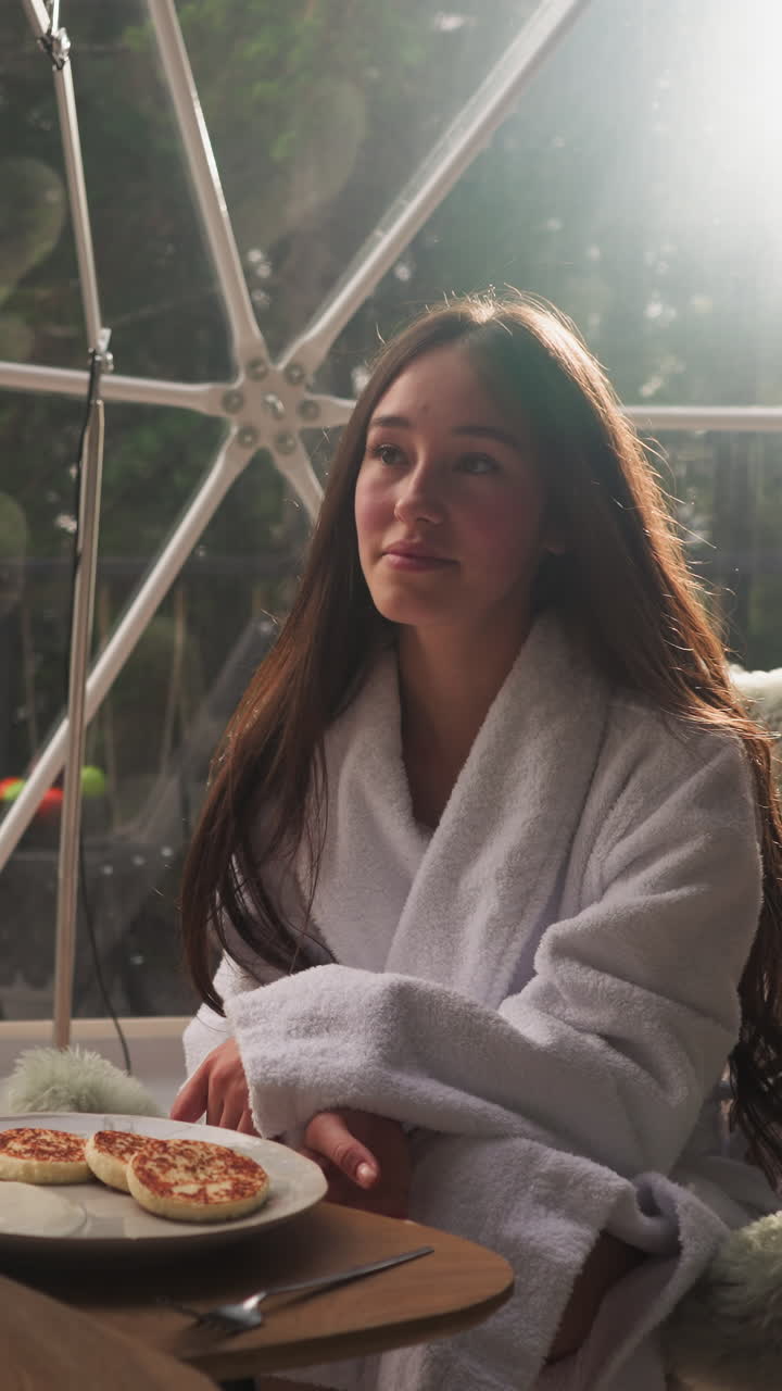 Woman sits at served table in round glamping house. Thoughtful brunette lady has pancakes for breakfast in tent with glass wall. Holiday at eco resort