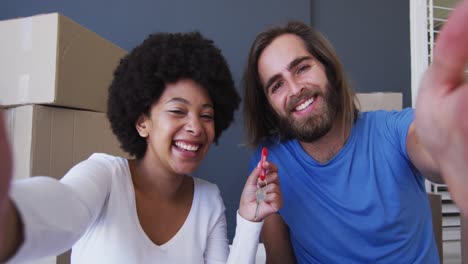 Portrait-of-mixed-race-couple-holding-keys-on-video-call-at-new-apartment-house