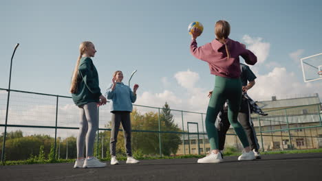 tres damas jugando al voleibol entre sí con el entrenador usando una camisa a cuadros alrededor de su cintura, cancha al aire libre con fondo de aro de baloncesto, valla y espacio verde