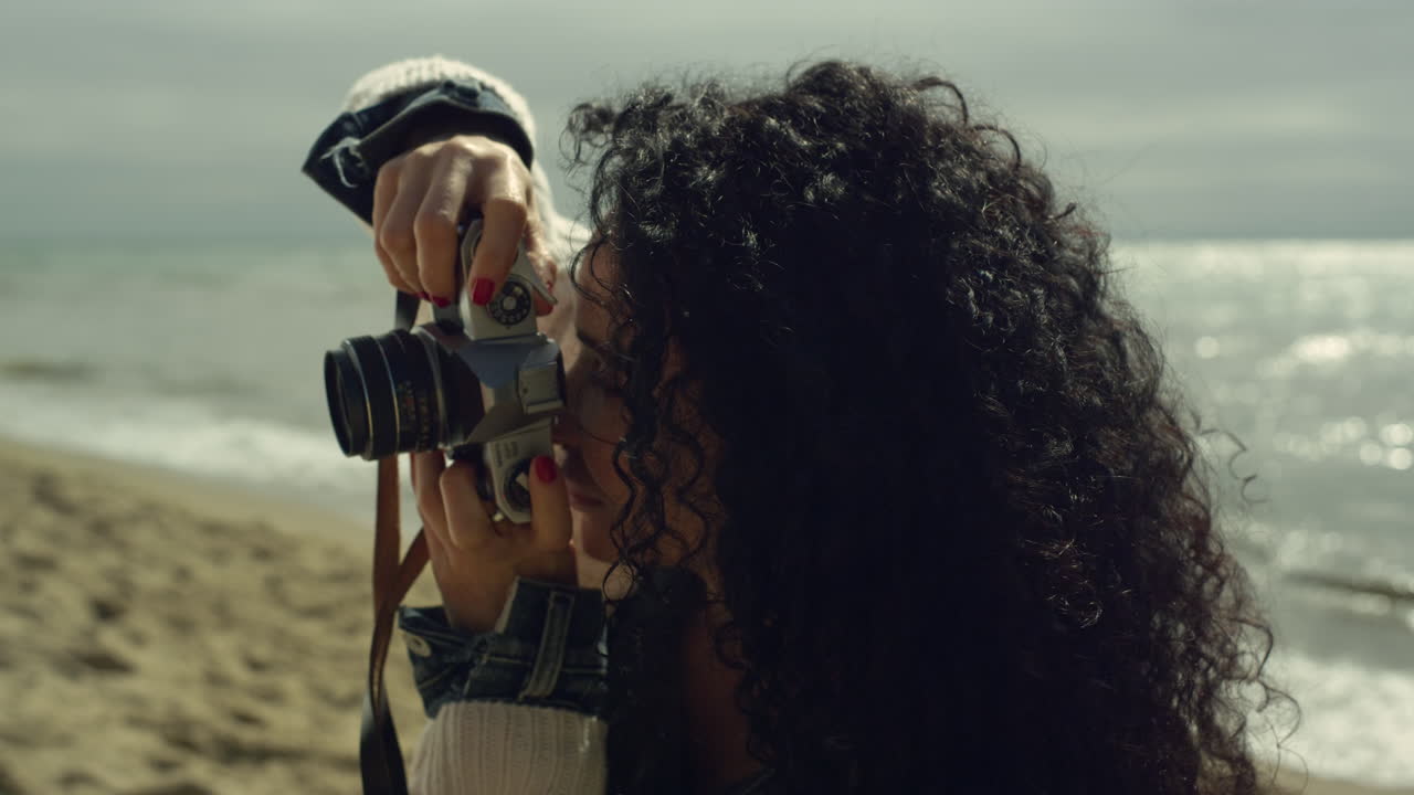 Hispanic girl photographing beach by calm sea. Curly hair woman taking photos.