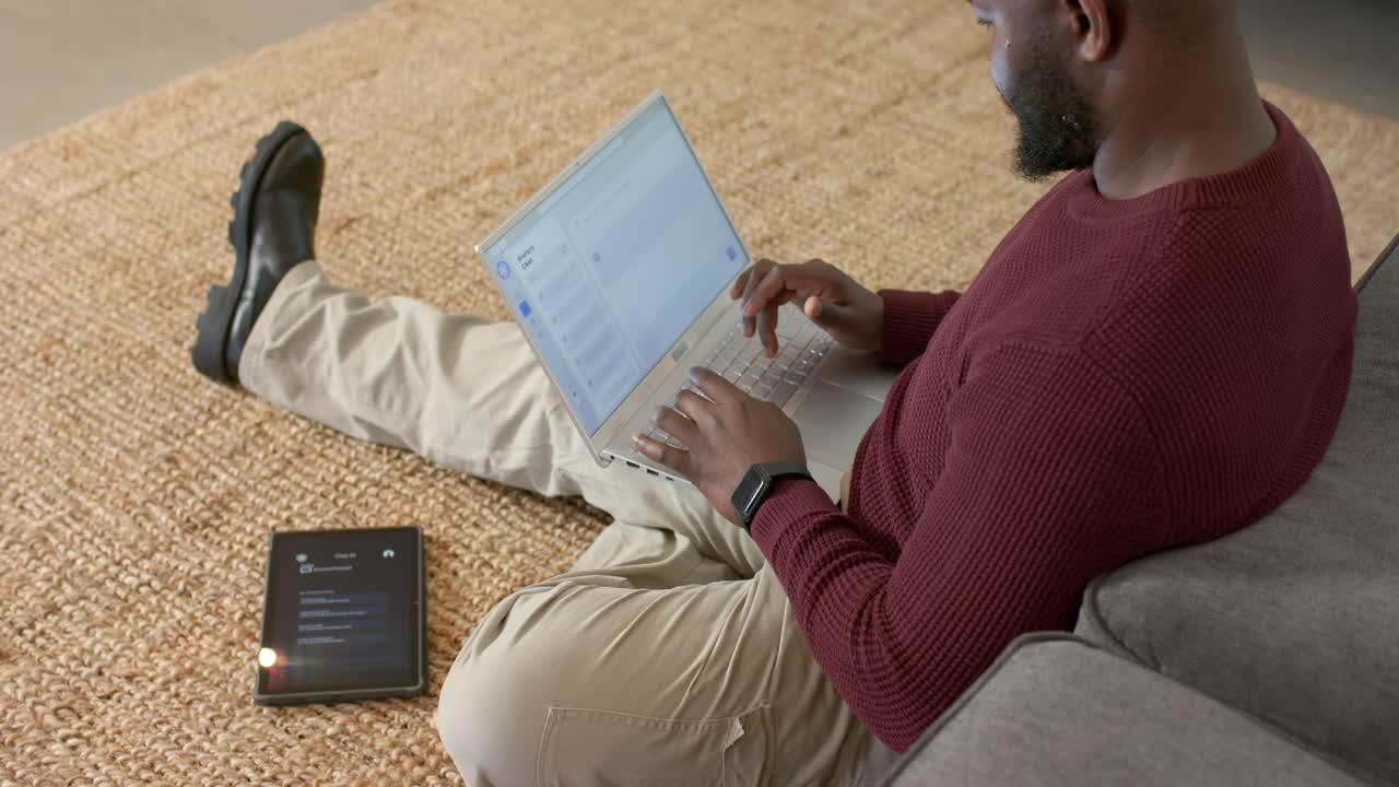 Mid adult African American man lifting laptop onto lap configuring files in living room with tablet