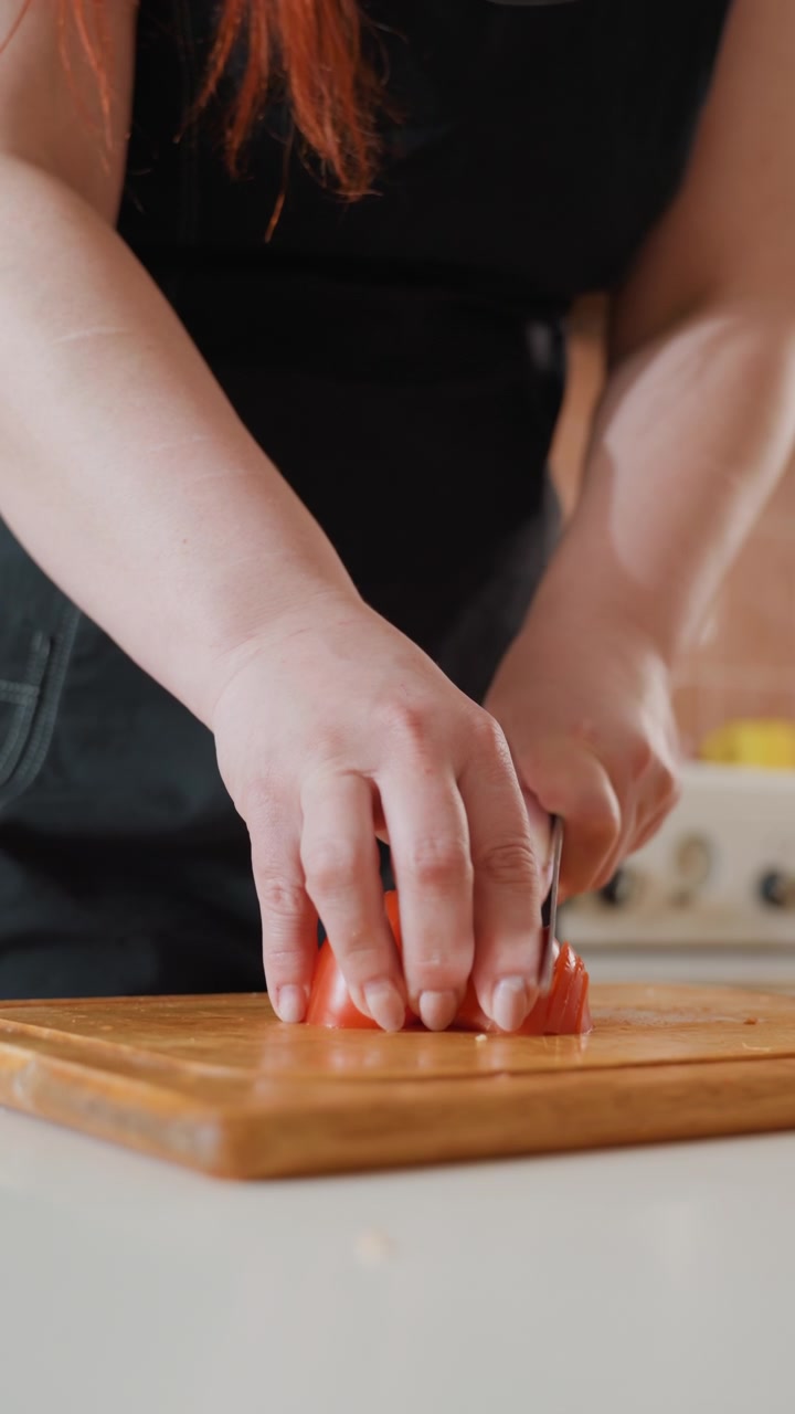 Primo piano di mani che tagliano pomodorini ciliegino su un tagliere di legno con un coltello piccolo, mostrando un movimento verso il basso, pezzi sparsi, succo fresco, in un ambiente caldo di cucina casalinga e con messa a fuoco ridotta.