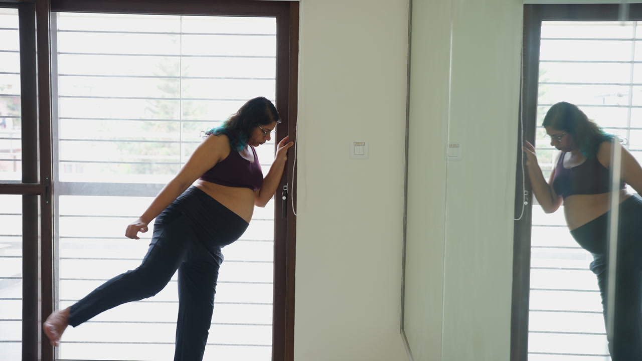 Indian female does hip rotation exercises in front of gym mirror in third trimester of pregnancy