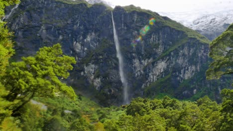 vasta cascada que cae por la montaña durante el día soleado rodeada de plantas y árboles verdes - glaciar rob roy, nueva zelanda