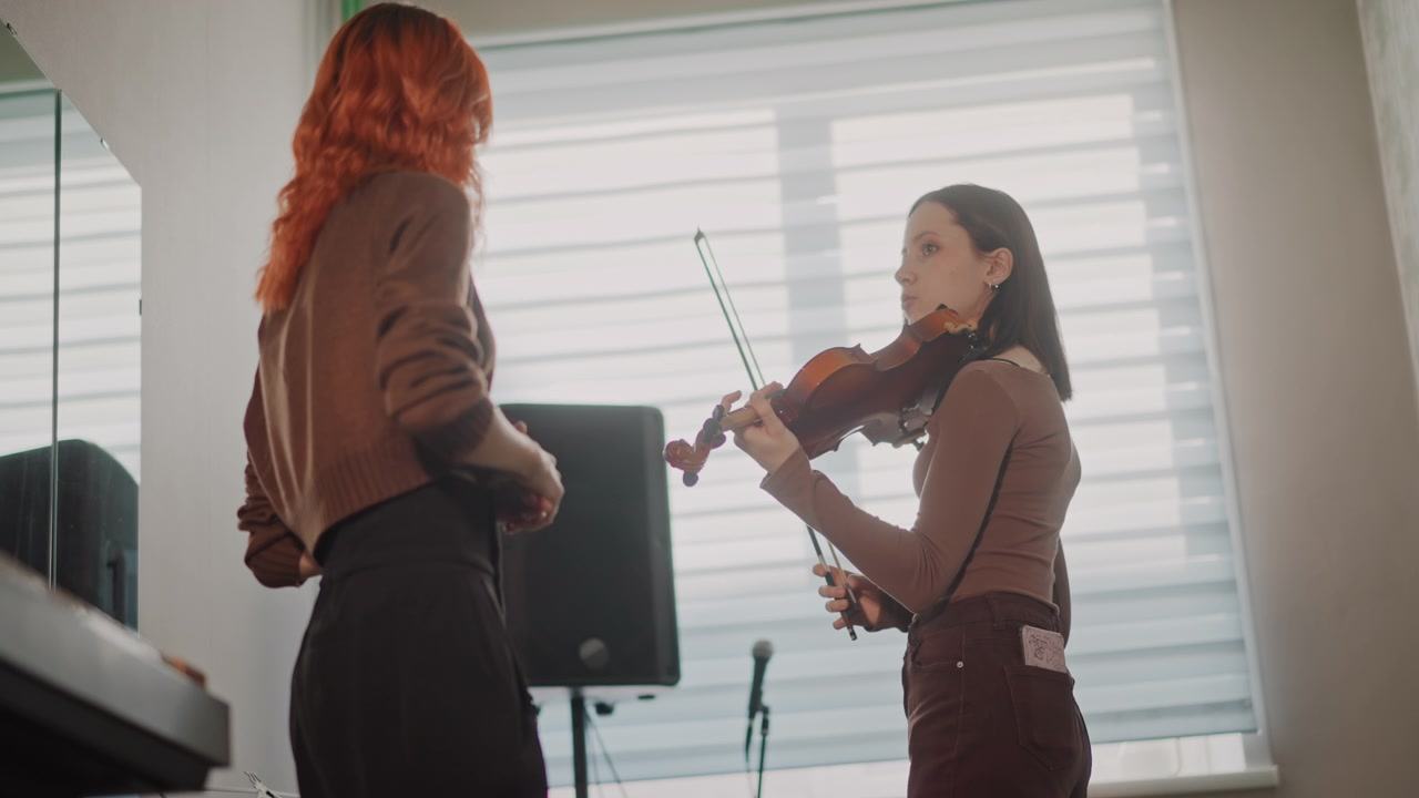 Female musicians practicing duet in bright studio with violin and bow, peer coaching and realtime feedback, speaker and mic setup, focused posture, collaborative energy and recital preparation vibe