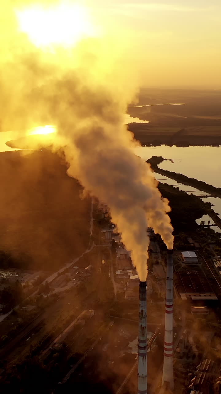Industrial plant near the river. Metallurgical factory with large pipes. Dirty emissions coming out of pipes into the air at sunset. Aerial view. Vertical video