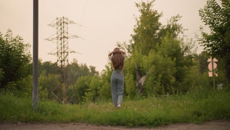 back view of young woman walking on grassy path with hands resting on head, surrounded by lush greenery, distant power lines above, and a car passing blurred in background