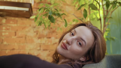 woman sits back and looks directly at camera while resting head on cushion in cozy room with warm brick wall and leafy plants, evoking calm mood and quiet confidence in relaxed natural setting