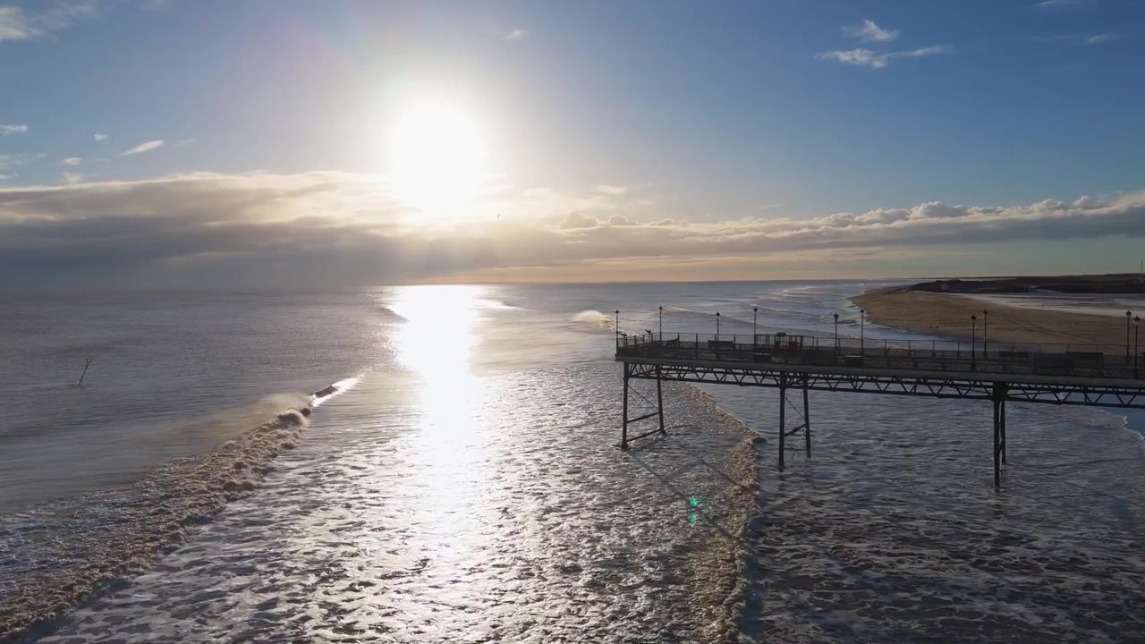 Il sole tramonta sotto l'orizzonte, inondando l'oceano di luce dorata. Le onde si infrangono dolcemente contro la base del molo di legno. Le persone passeggiano lungo il molo, con espressioni rilassate e contente