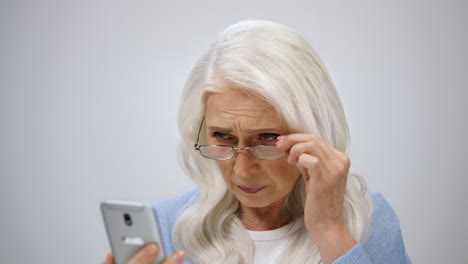 old woman fixing glasses in modern studio. annoyed lady looking to phone screen.