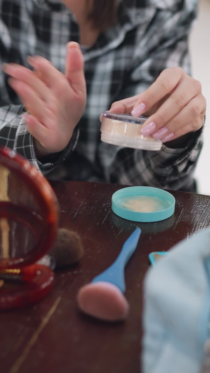 여성이 루스 파우더로 화장을 준비하고, 거울 옆에서 캐주얼한 체크무늬 셔츠를 입고 있어요. 테이블 위에는 브러시와 파우치가 있고, 손은 집중하여 병을 두드리고 있네요. 따뜻한 자연광과 친근한 가정적인 분위기가 느껴지는 자연스러운 아침 일상이에요.