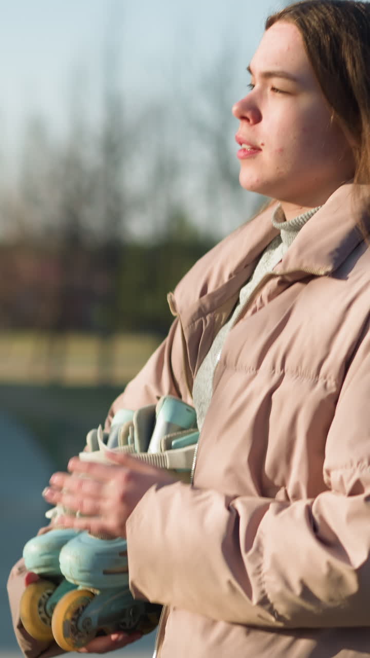 una chica lleva una patineta mientras camina por un parque en un día soleado. está vestida con una chaqueta de melocotón y una camisa interior gris, con la boca ligeramente abierta mientras mira hacia abajo