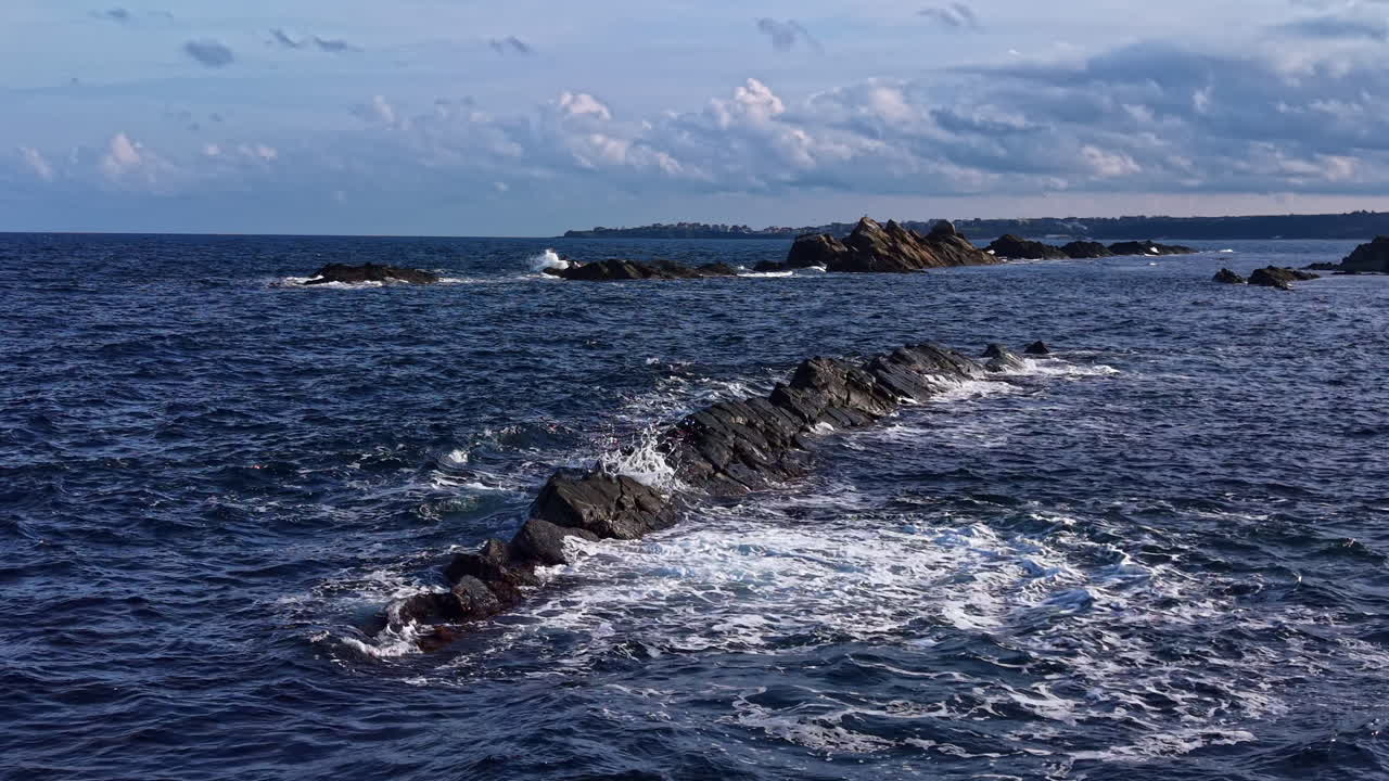Garis pantai berbatu di bawah langit tenang dengan ombak lembut memercik ke bebatuan di dekat pantai saat matahari terbenam