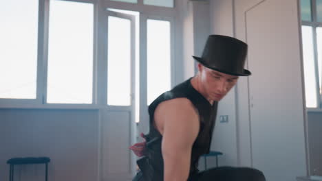 young man in black vest and hat kneels on floor mid dance with fluid arm gesture studio windows and side mirror framing reflective silhouette moody light and smoke accent for dramatic movement