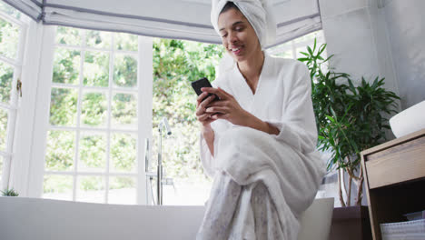 mixed race woman sitting on bathtub and using her smartphone in bathroom