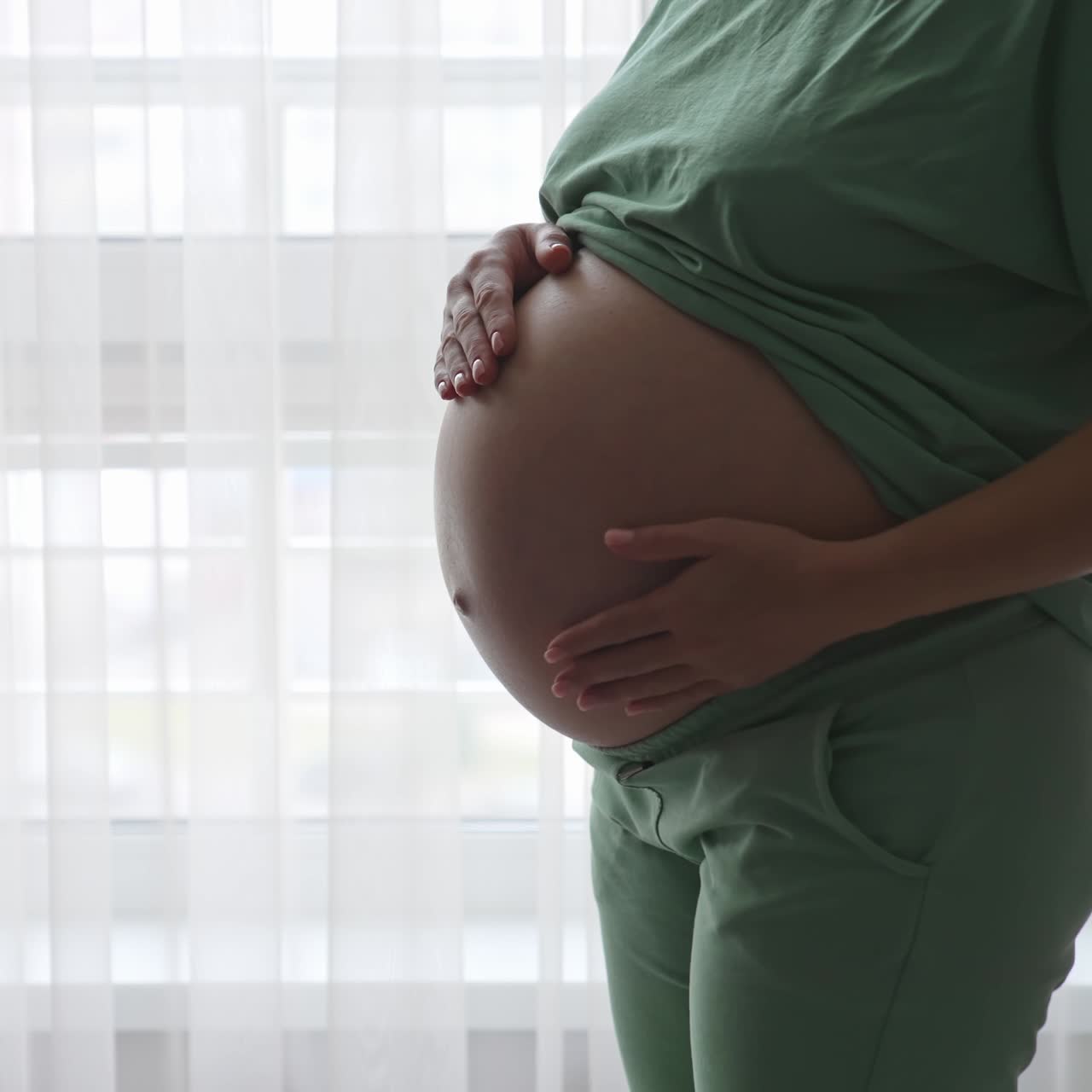 Futura mamma in piedi alla finestra che tocca delicatamente la sua pancia. La donna in attesa, in abiti comodi, accarezza amorevolmente la sua pancia.