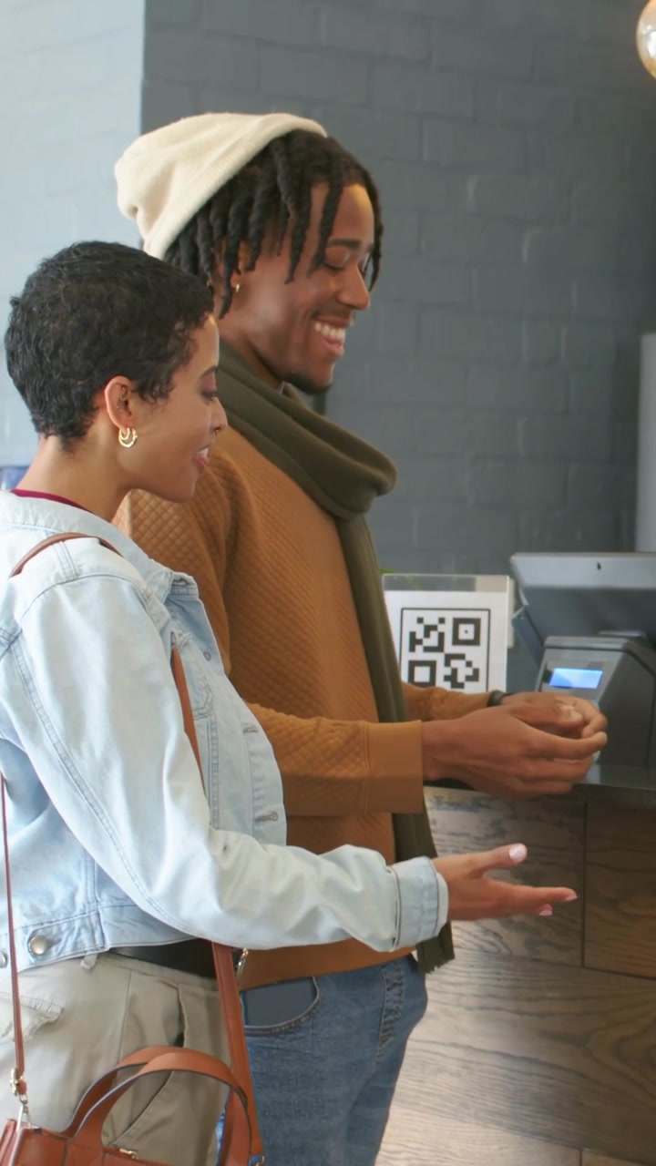 Vertical video: Accepting man taking coffees at cafe counter after scanning QR, in beanie, scarf