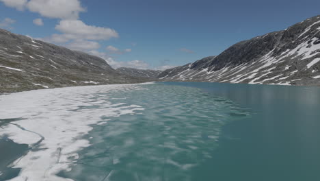 um drone em câmera lenta voando perto dos flocos de gelo no lago langvatnet, na noruega, perto do strynefjellsveg em um dia ensolarado, cercado por rochas brilhantes e montanhas.