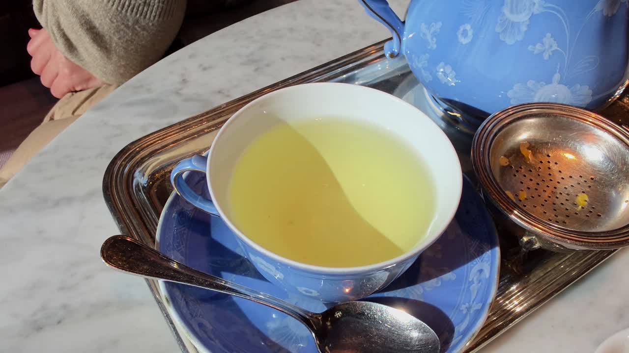 Primo piano di una mano femminile con manicure rossa che solleva una tazza di porcellana blu piena di tisana gialla da un vassoio d'argento su un tavolo di marmo