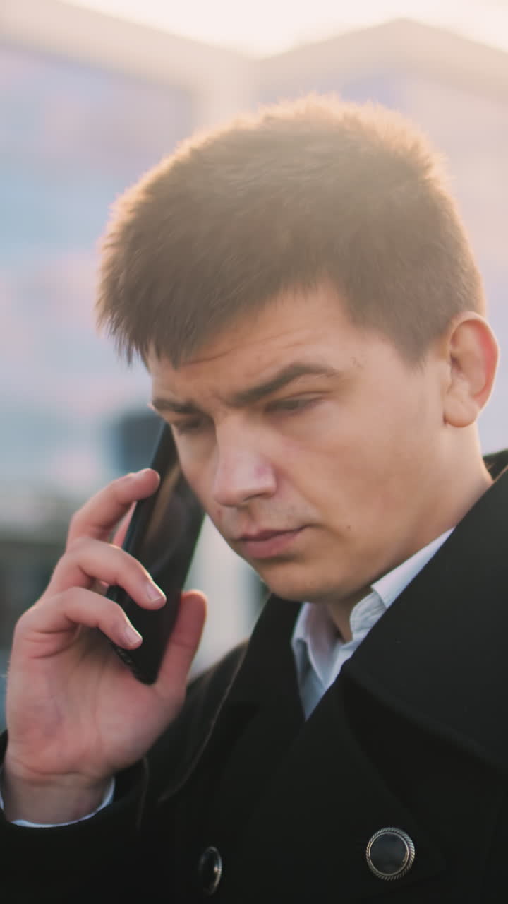 검은 코트를 입은 남자가 전화를 계속하는 동안  아래로 전화를 조정하고 동의로 고개를 끄덕인다. 배경에는 현대적인 유리 건물, 주차된 자동차, 따뜻한 빛이 있습니다.