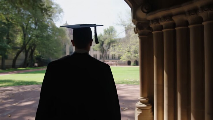 Graduate In Cap And Gown Walking Through Collegiate Archway Into Sunlit Quad, Back View Of Student