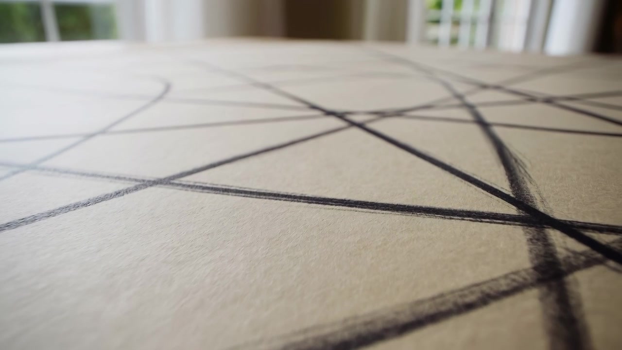 Close perspective reveals dark intersecting lines on pale textured material as camera movement changes framing, with soft window light and blurred background creating calm geometric surface imagery.