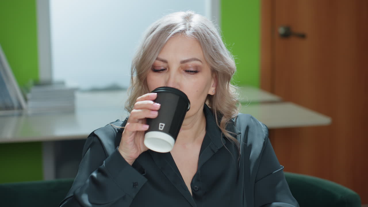 Office assistant pauses while working on laptop to sip coffee before continuing typing, seated in modern office with folders, documents, and natural light creating calm focused work environment