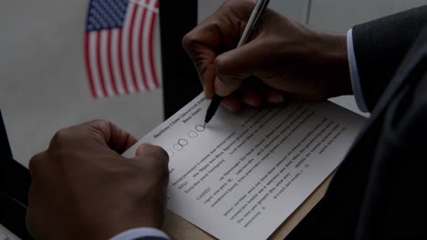 a person is shown writing on a document with a pen, with an american flag visible in the background