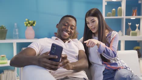 Happy-African-American-Young-man-and-Caucasian-Young-woman-using-phone-together-at-home-ordering-online.