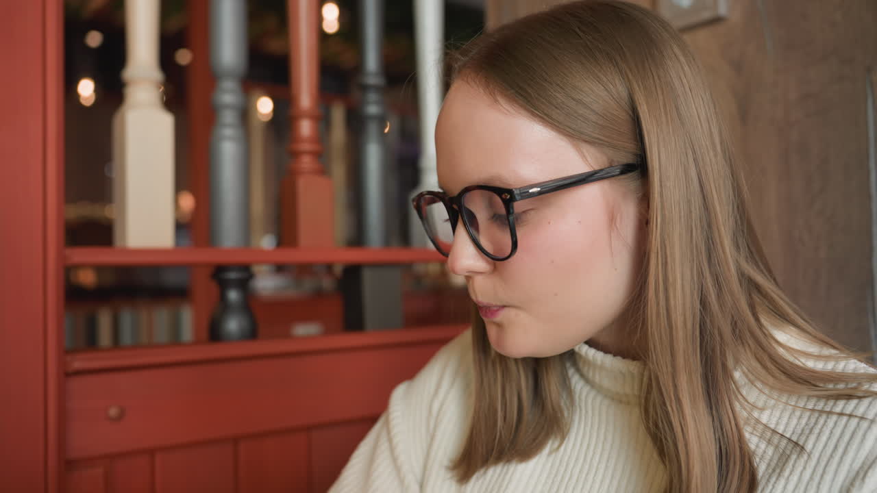 Realtor in white sweater and glasses sipping latte in cozy cafe with visible wall socket and blurred bar lights in background creating warm ambiance and calm indoor atmosphere