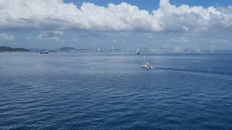 a catamaran sails on calm ocean waters with the skyline of panama city visible in the background under a partly cloudy sky.