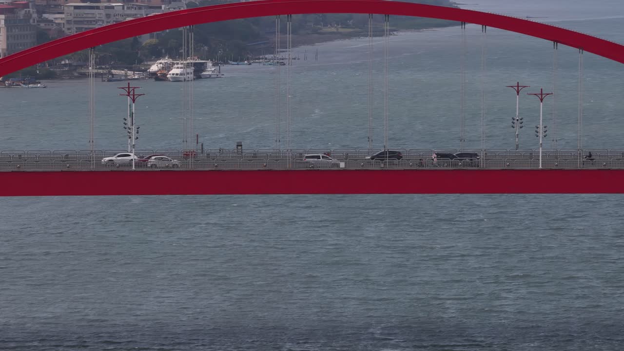 Vista aérea de vehículos circulando por un moderno puente de arco rojo sobre una gran masa de agua con viento, con una ciudad costera visible al fondo conectando la infraestructura urbana