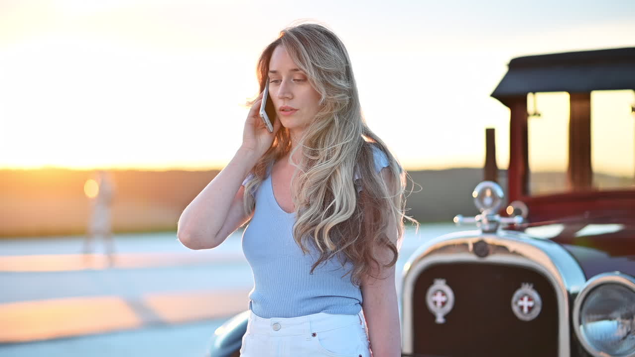 Stylish woman standing beside a vintage car while talking on her phone during sunset