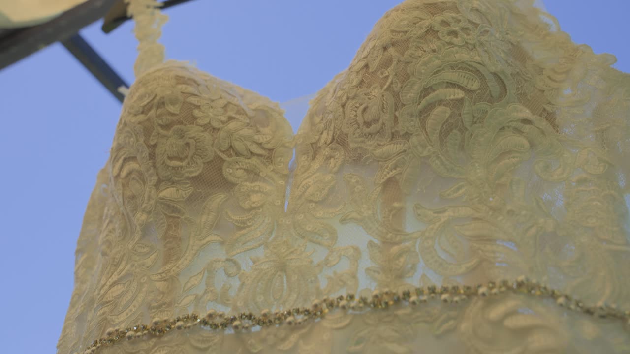 Una carrellata dal basso di un bellissimo abito da sposa bianco caratterizzato da un intricato pizzo floreale, una cintura gioiello scintillante e spalline delicate, appeso all'aperto in una giornata limpida.