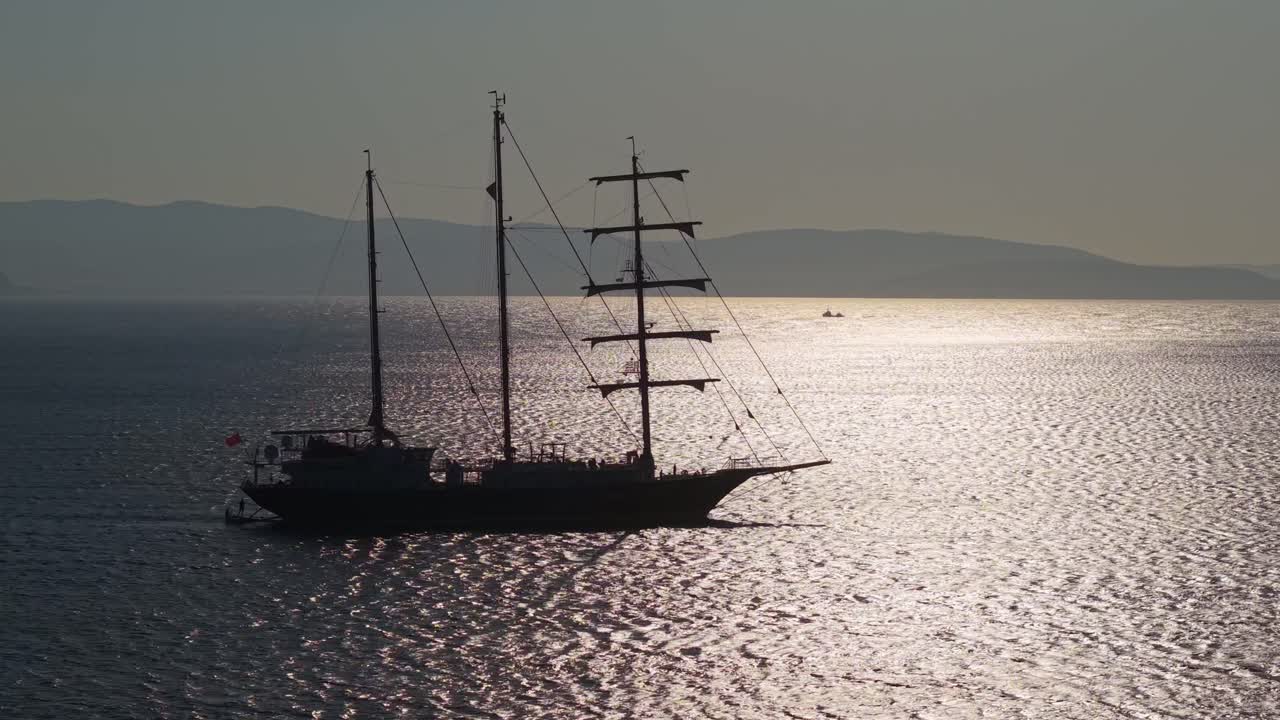A beautiful three-masted sailboat glides across the Aegean Sea as the sun sparkles and reflects on the calm sea surface, creating a serene maritime scene