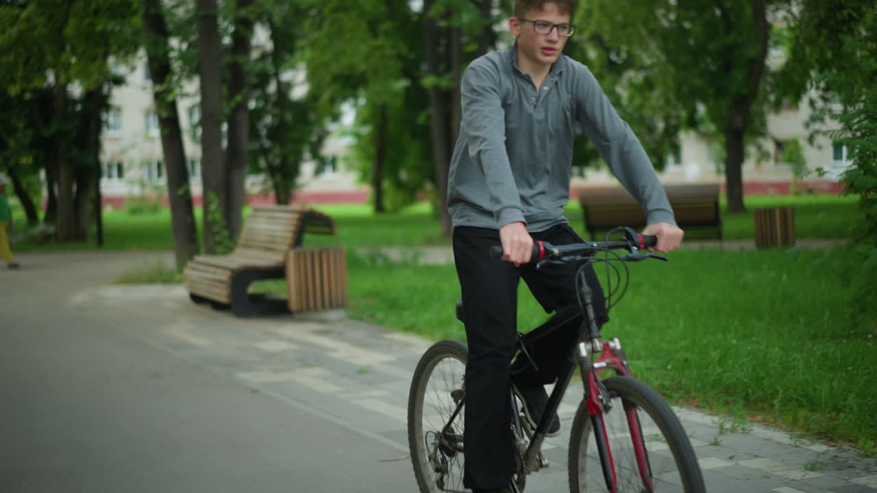 회색 과 검은색 바지를 입은 소년은 자전거를 타고 푸른 나무, 나무 벤치, 그리고 평화로운 도시 환경의 배경에 있는 주거 건물로 둘러싸인 공원 길을 따라 달린다.