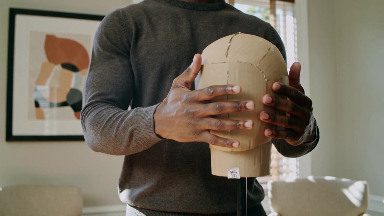 Adjusting man aligning cardboard head form at home, smoothing seams for camera, wearing sweater