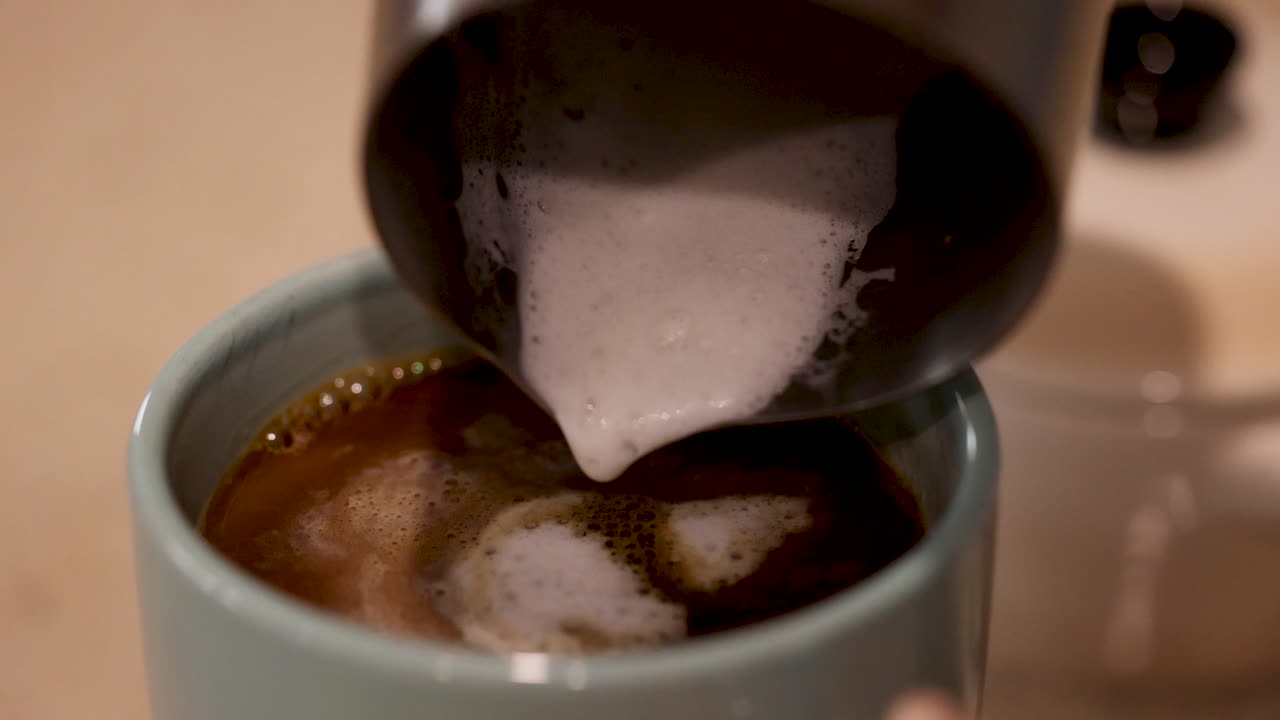 Milk is poured into a steaming cup of coffee, creating swirling patterns. Warm lighting enhances the cozy atmosphere