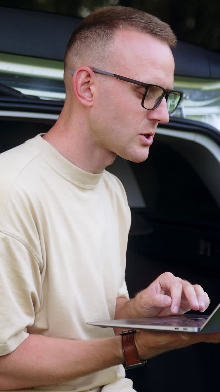 Mid-aged freelancer focused on laptop. Man works sitting in car trunk in the forest. Vertical video