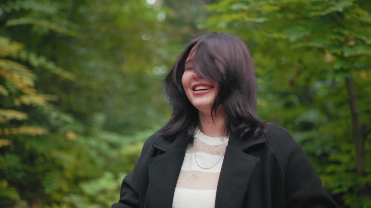푸른 숲에서 머리카락의 먼지를 털어내는 차분한 여성, 부드럽게 흐릿한 배경, 부드러운 미소, 캐주얼 코트, 장난스러운 나뭇잎 던지기 후 편안한 가을 산책 중 자기 관리를 하는 자연스러운 순간, 클로즈업 시점