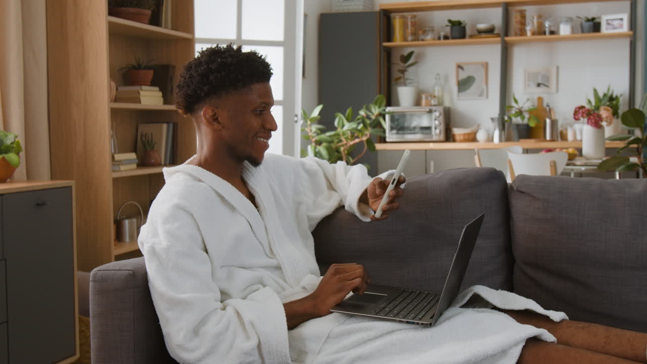 Man in robe using laptop and smartphone on sofa at home