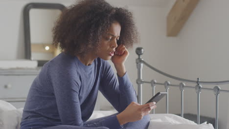 Unhappy-Woman-Wearing-Pyjamas-Sitting-On-Bed-With-Mobile-Phone-Being-Bullied-Online-Via-Social-Media