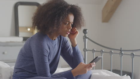 unhappy woman wearing pyjamas sitting on bed with mobile phone being bullied online via social media