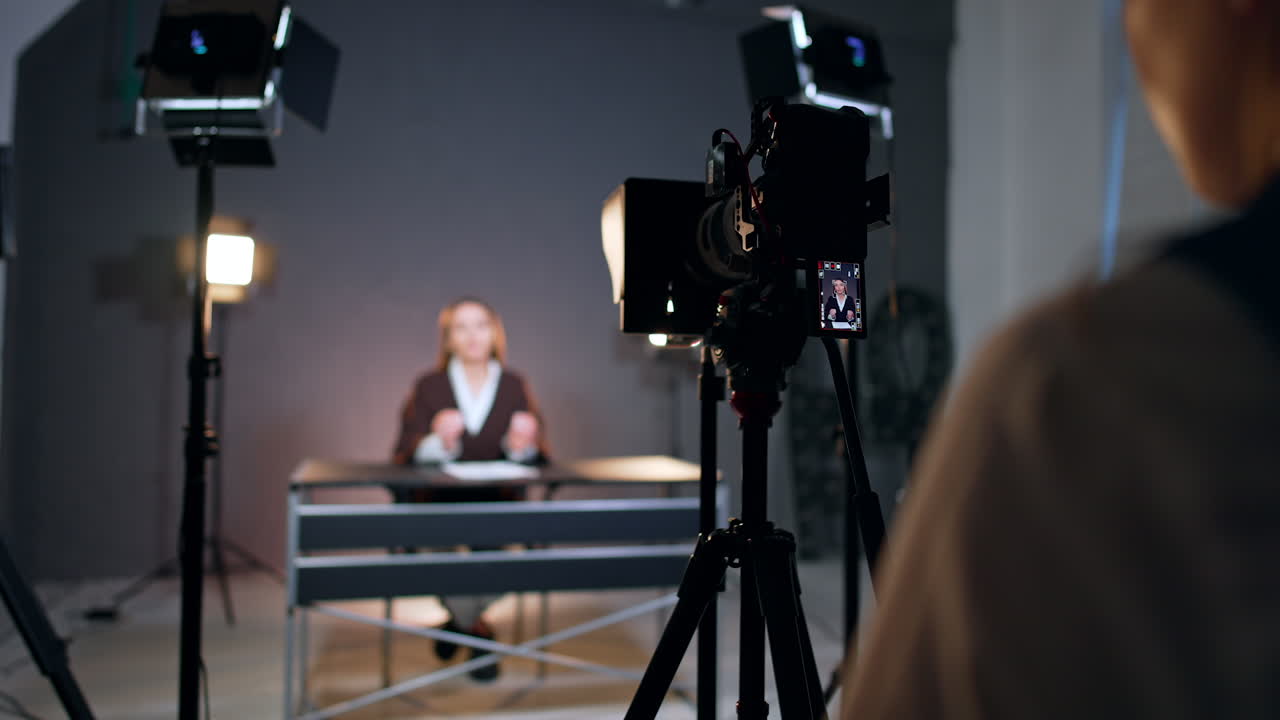 Unrecognized woman stands backstage behind the camera. Woman sits at desk at blurred backdrop talking and waving hands